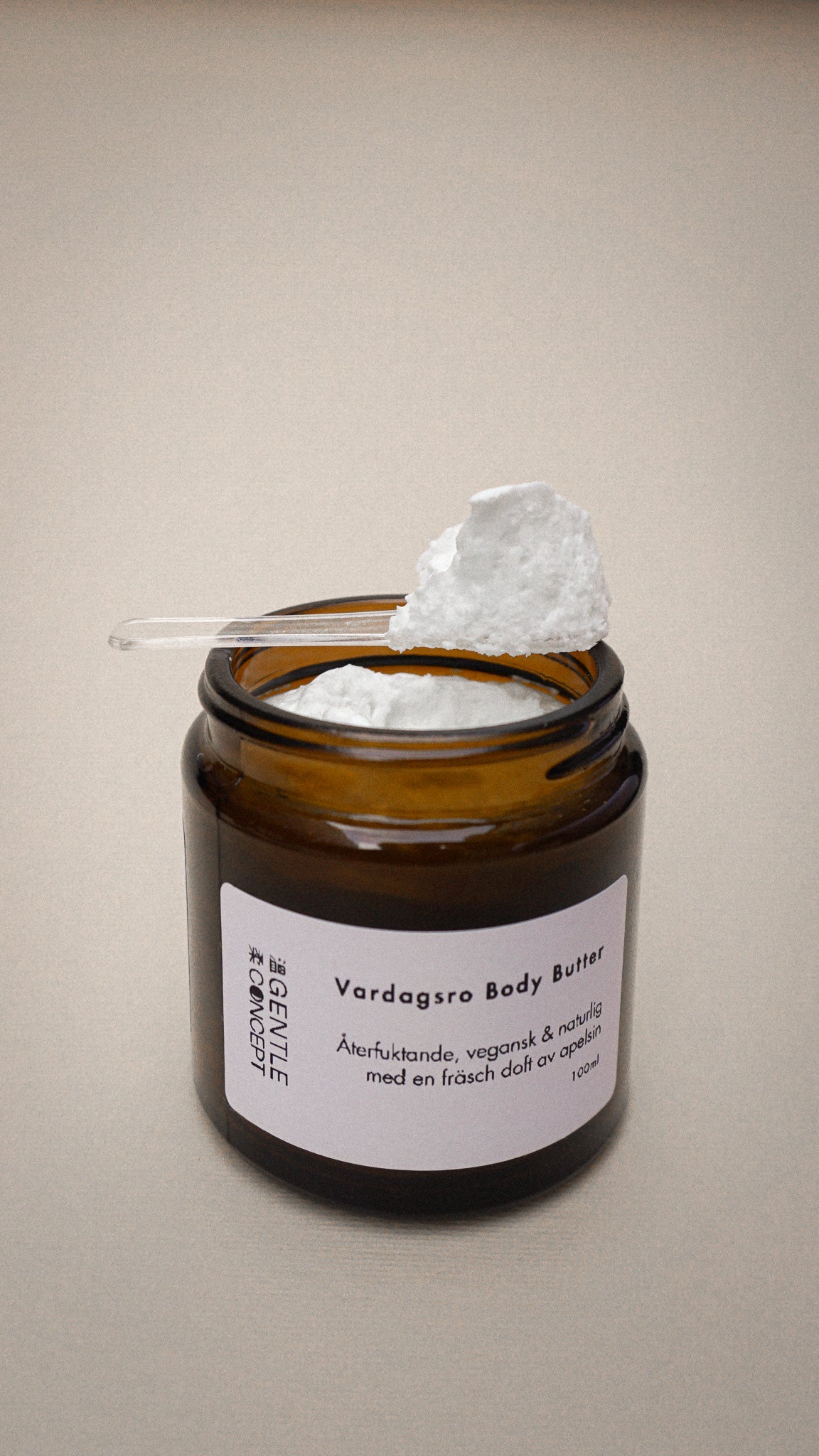 En bild på en glasburk med vårdande hudkräm där produktens textur visas på en spatel. Bakgrunden är neutral med beige färg och burken har en beskrivande etikett på sig där det står Vardagsro Body Butter: Återfuktande, vegansk och naturlig med en fräsch doft av apelsin.