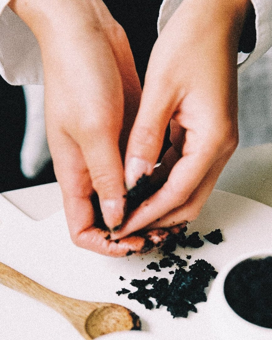 Close-up of hands holding some coffee-based body scrub, with a small wooden spoon beside it on a clean background.